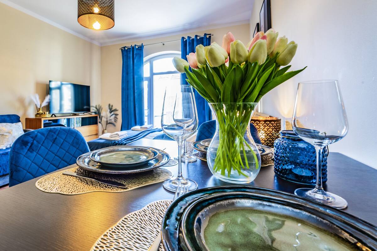 Un área de comedor con una mesa preparada para dos personas, un sofá azul, un televisor y una ventana con cortinas.
