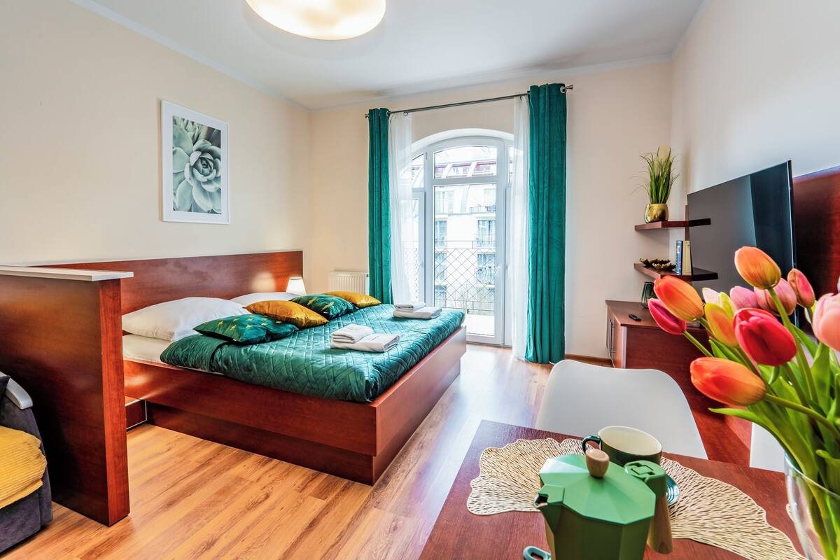 Habitación de hotel moderna con una cama de madera, un escritorio pequeño, televisión y vistas a la ciudad desde una ventana.