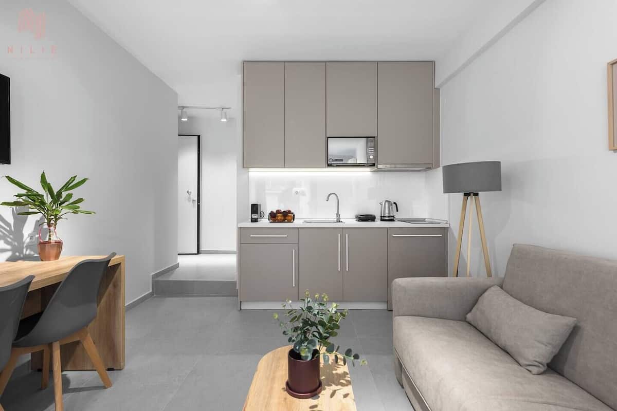 A modern kitchen with grey cabinets, a microwave, and a coffee maker. There is a dining area with a wooden table and chairs, and a grey sofa in the living area.