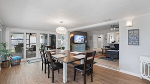 A spacious living area with a dining table, chairs, and a television. There is a kitchen in the background, and a hallway leading to another room.