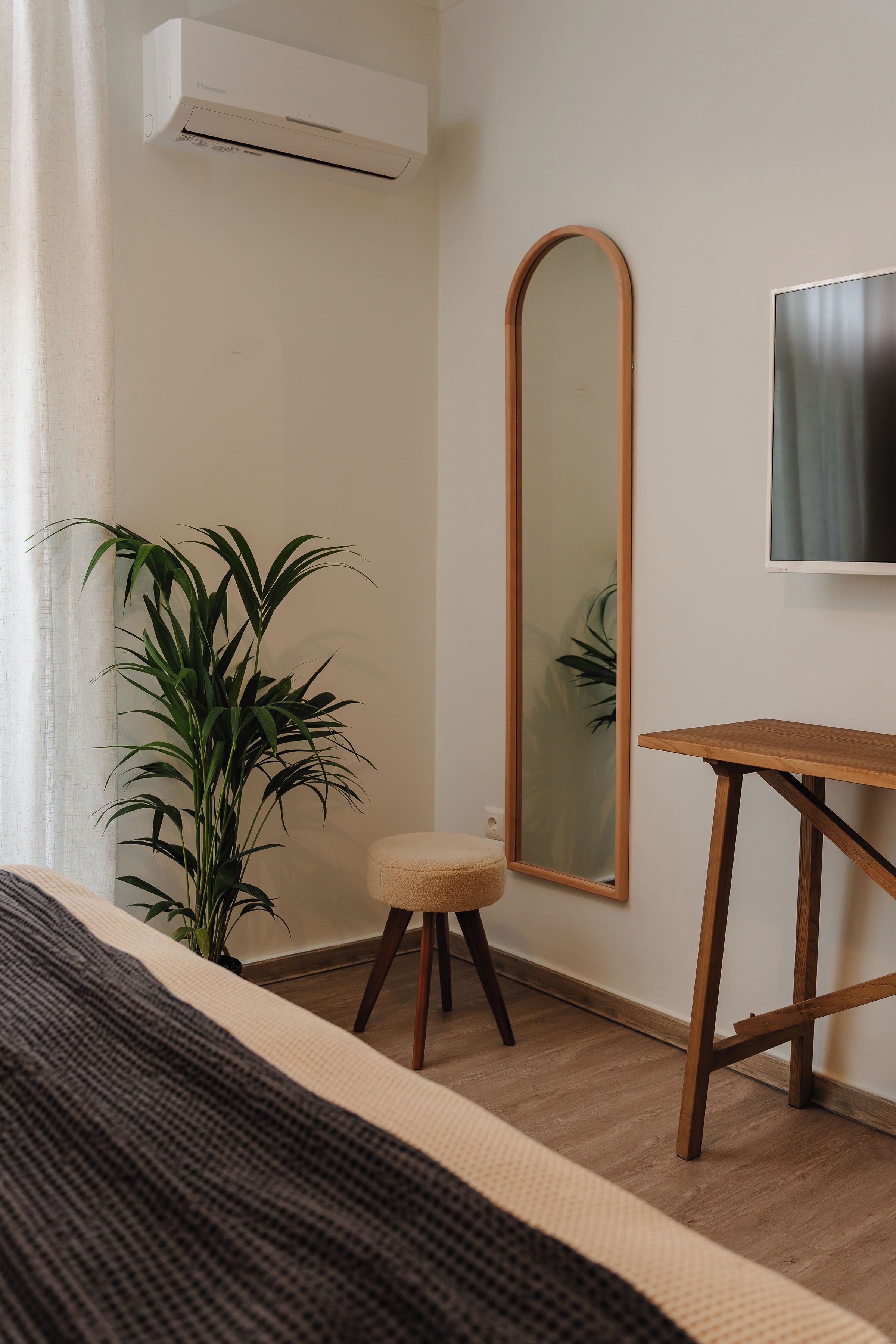 Un dormitorio con cama, mesita de noche de madera, espejo, planta en maceta y equipo de aire acondicionado.