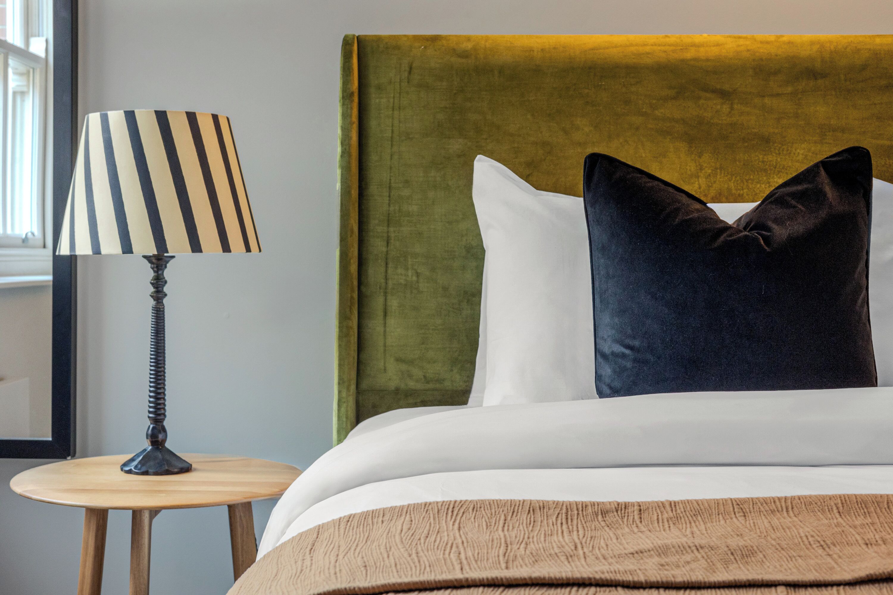 A neatly made bed with a green headboard, a striped lamp on a wooden side table, and a white pillow.