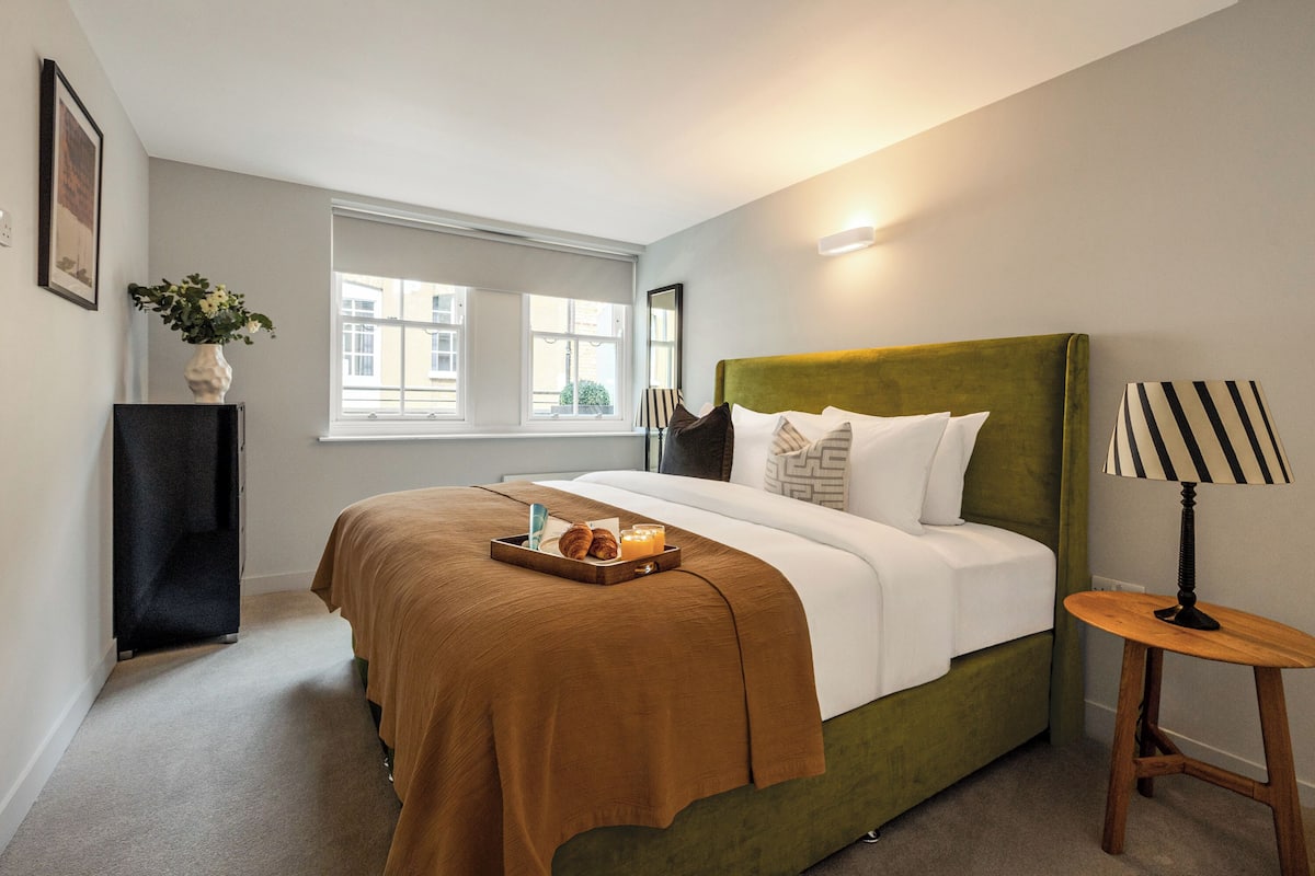 A hotel room with a green upholstered bed, a bedside table with a lamp, a tray with croissants and juice, and a window with a view of buildings.