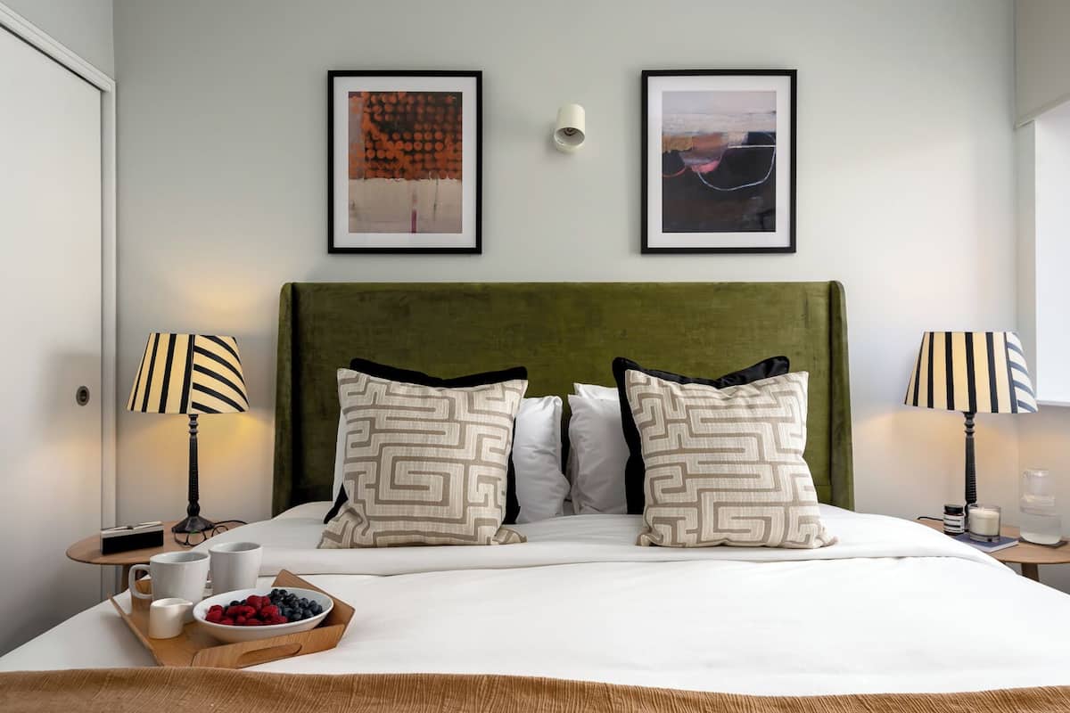 A neatly made bed with a green headboard, two bedside tables with lamps, and two framed artworks on the wall.