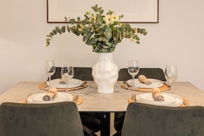 A dining table set for four with white plates, folded napkins, and wine glasses. A white vase with green foliage is centered on the table.