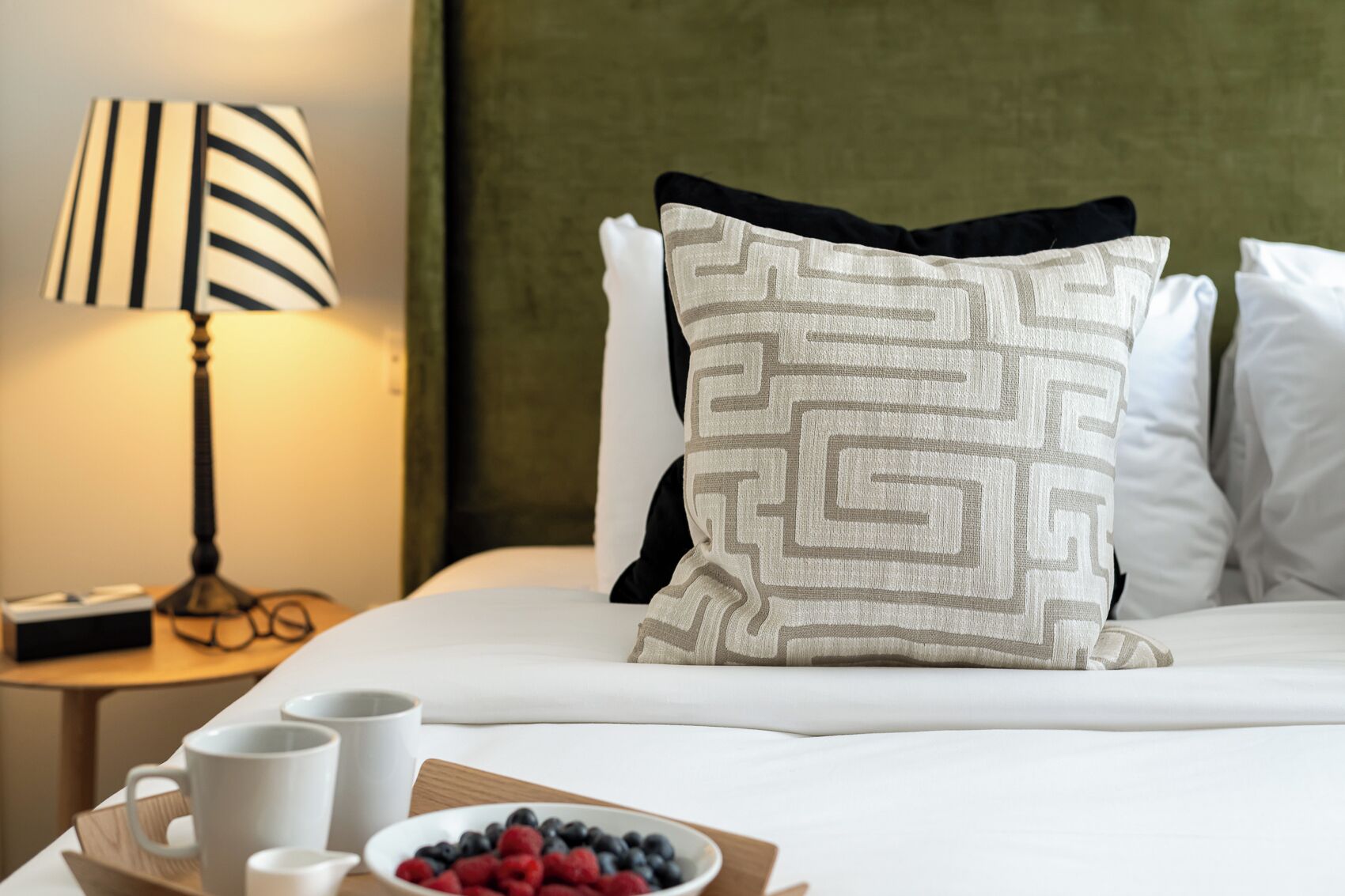 Una cama bien hecha con un cojín estampado, una mesita de noche con una lámpara, y un bol de bayas.