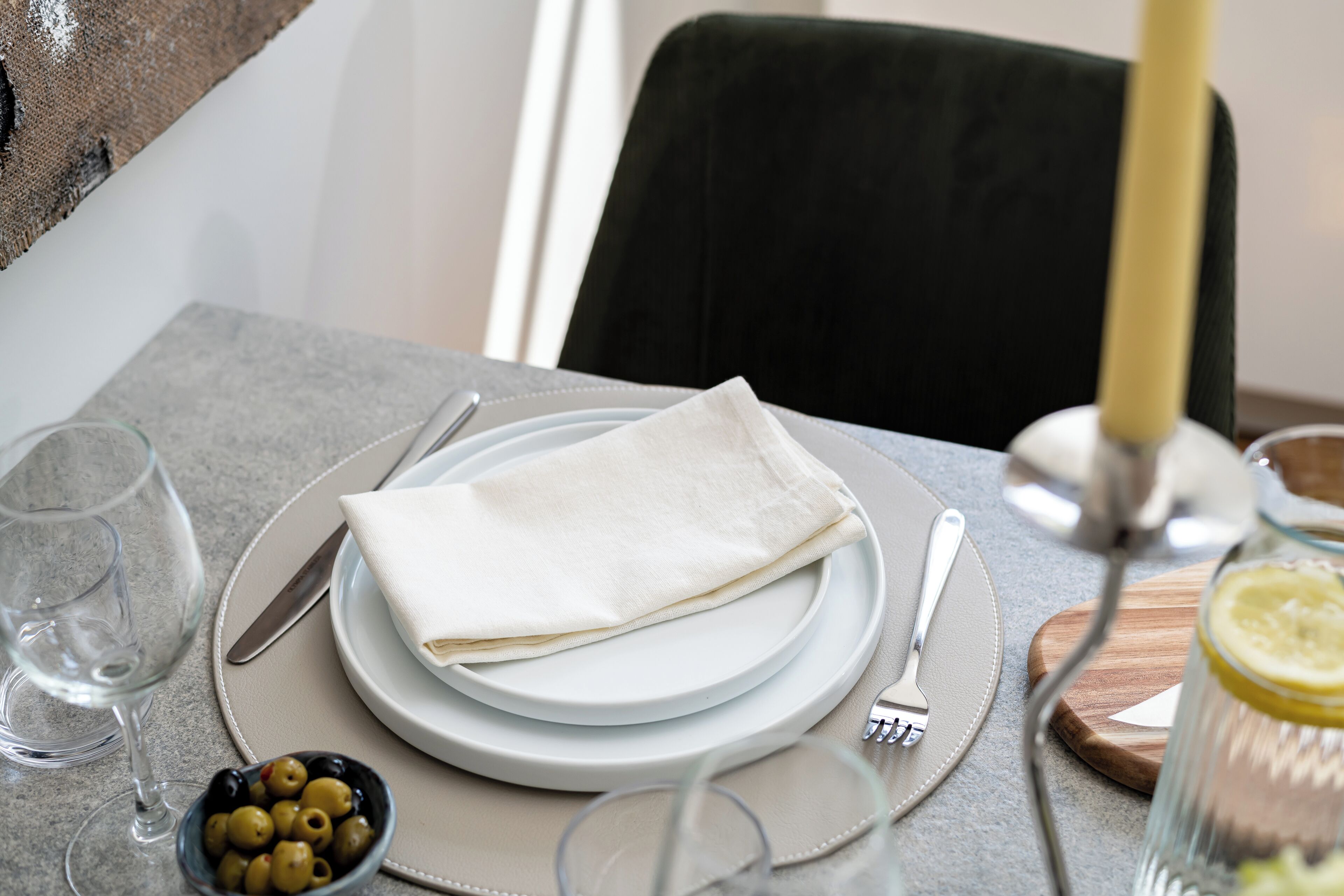 Una mesa de comedor dispuesta con platos, cubiertos, un vaso de agua, un cuenco de aceitunas y un limón en un tarro de cristal.