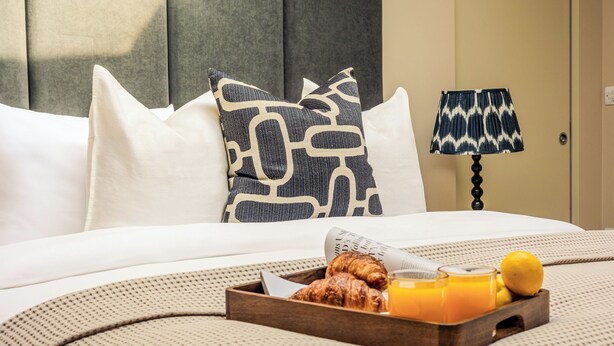 A neatly made bed with white linens, a tray of croissants, orange juice, and a lemon. - Your Apartment - Liverpool Street (London)