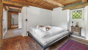 Une chambre avec un plafond en bois, un lit recouvert de draps blancs, une table de chevet en bois et une fenêtre donnant sur un paysage verdoyant.