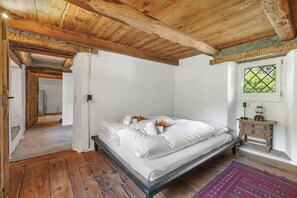 Ein Schlafzimmer mit Holzboden, einem Bett mit weißen Leinen, einem Nachttisch aus Holz und einem Fenster mit Blick auf Grünflächen.