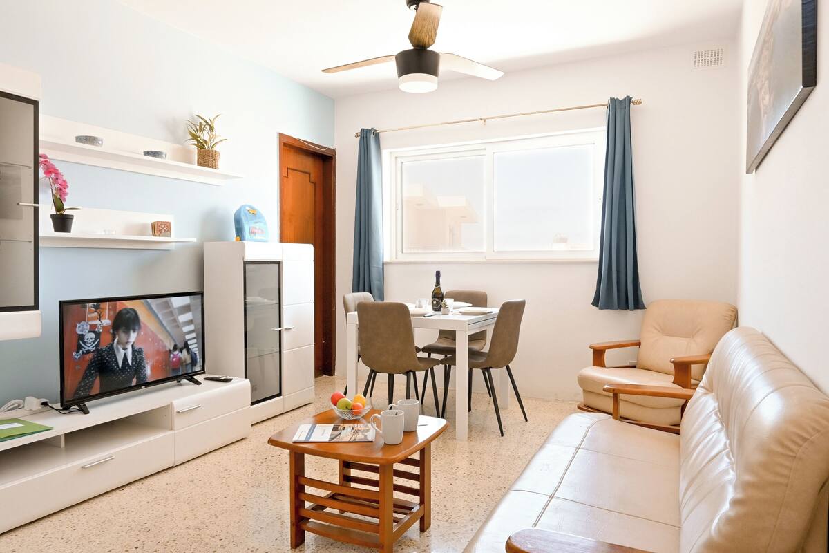 A modern living room with a flat-screen TV, a dining table with chairs, a sofa, and a ceiling fan.