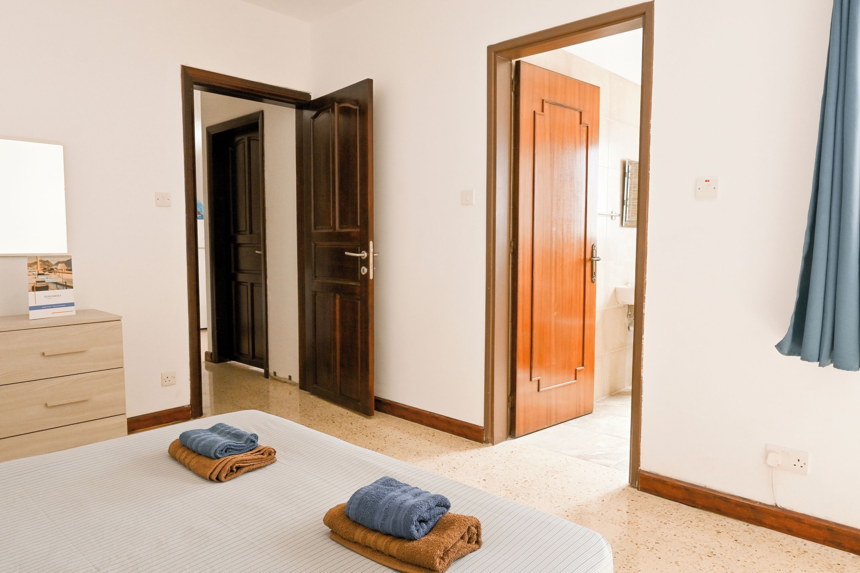 Habitación de hotel con cama, puerta de madera, baño visible a través de una puerta abierta y una cortina azul.