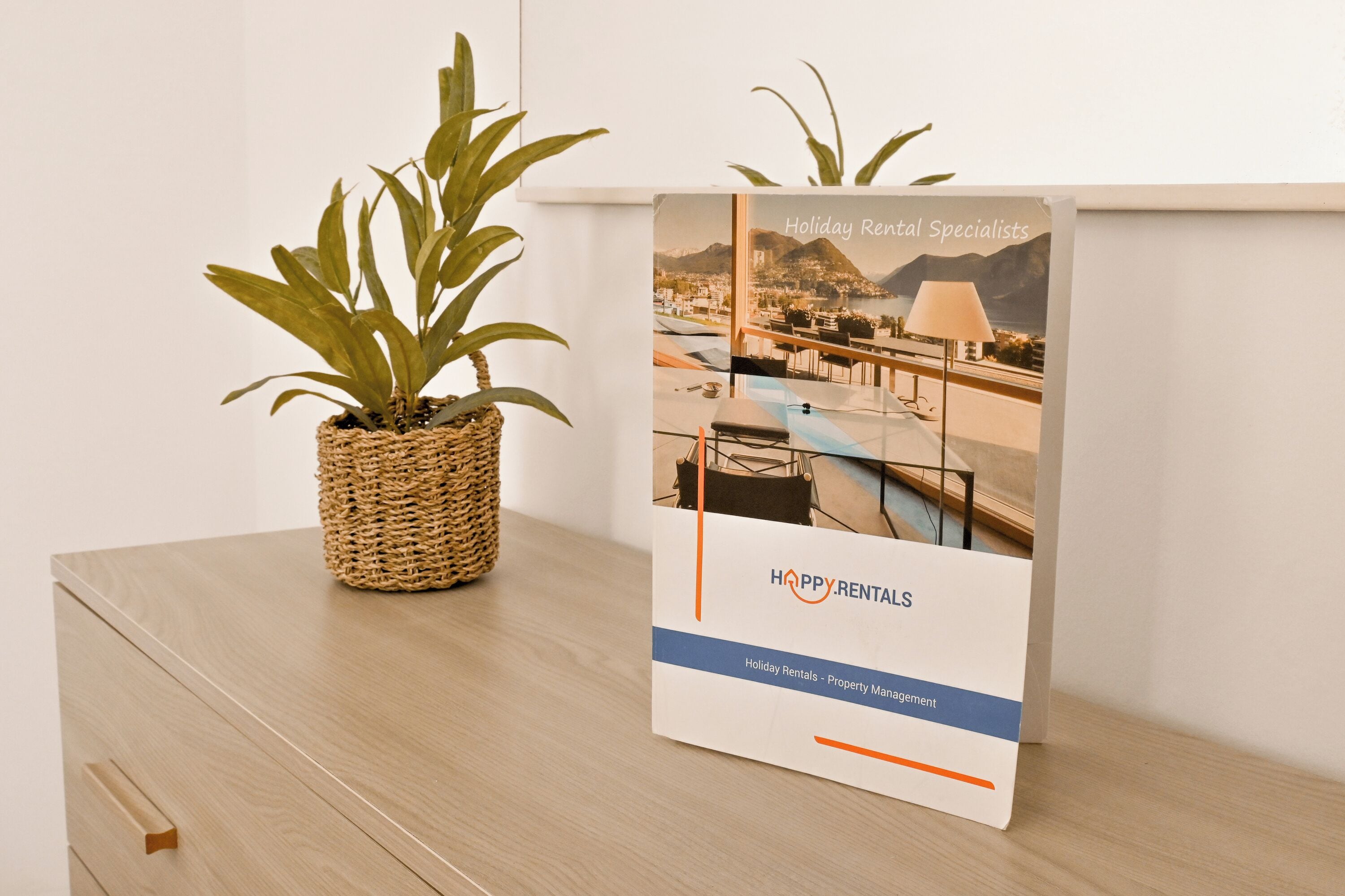 Un folleto con el título 'Holiday Rental Specialists' que muestra una vista panorámica de la costa, una planta en mimbre y una mesa de madera.