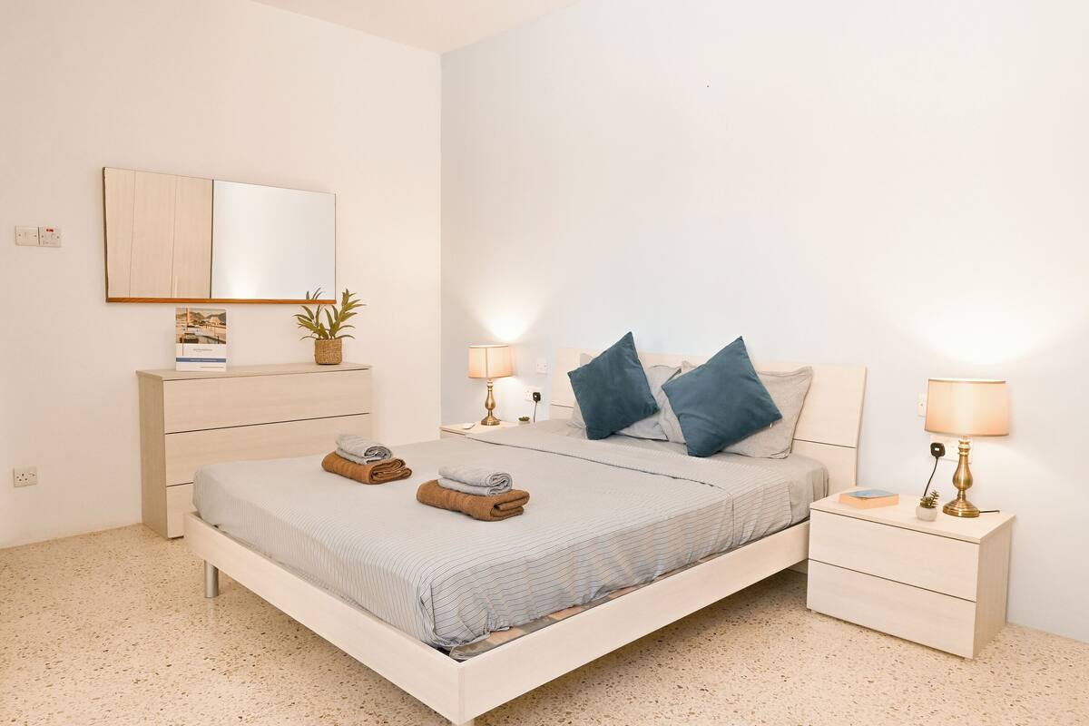 A modern bedroom with a bed, bedside tables, a nightstand, a mirror, and a wall-mounted shelf.