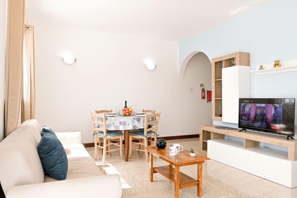 A living room with a sofa, a coffee table, a dining table with chairs, a TV on a wooden stand, and a bookshelf.