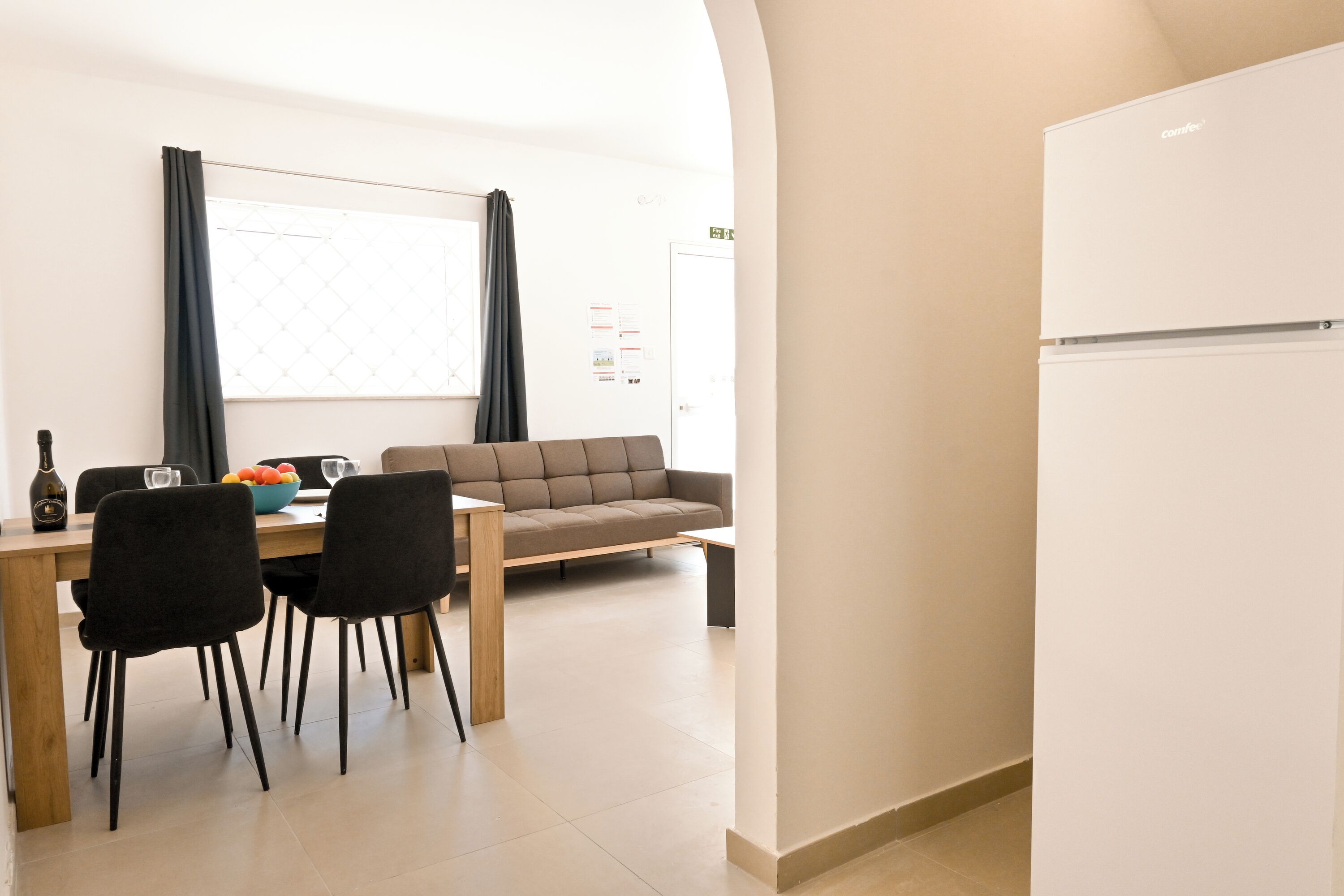 Ein modernes Wohnzimmer mit einer Couch, einem Esstisch und Stühlen. Ein weißer Kühlschrank und ein Fenster mit Vorhängen sind ebenfalls vorhanden.