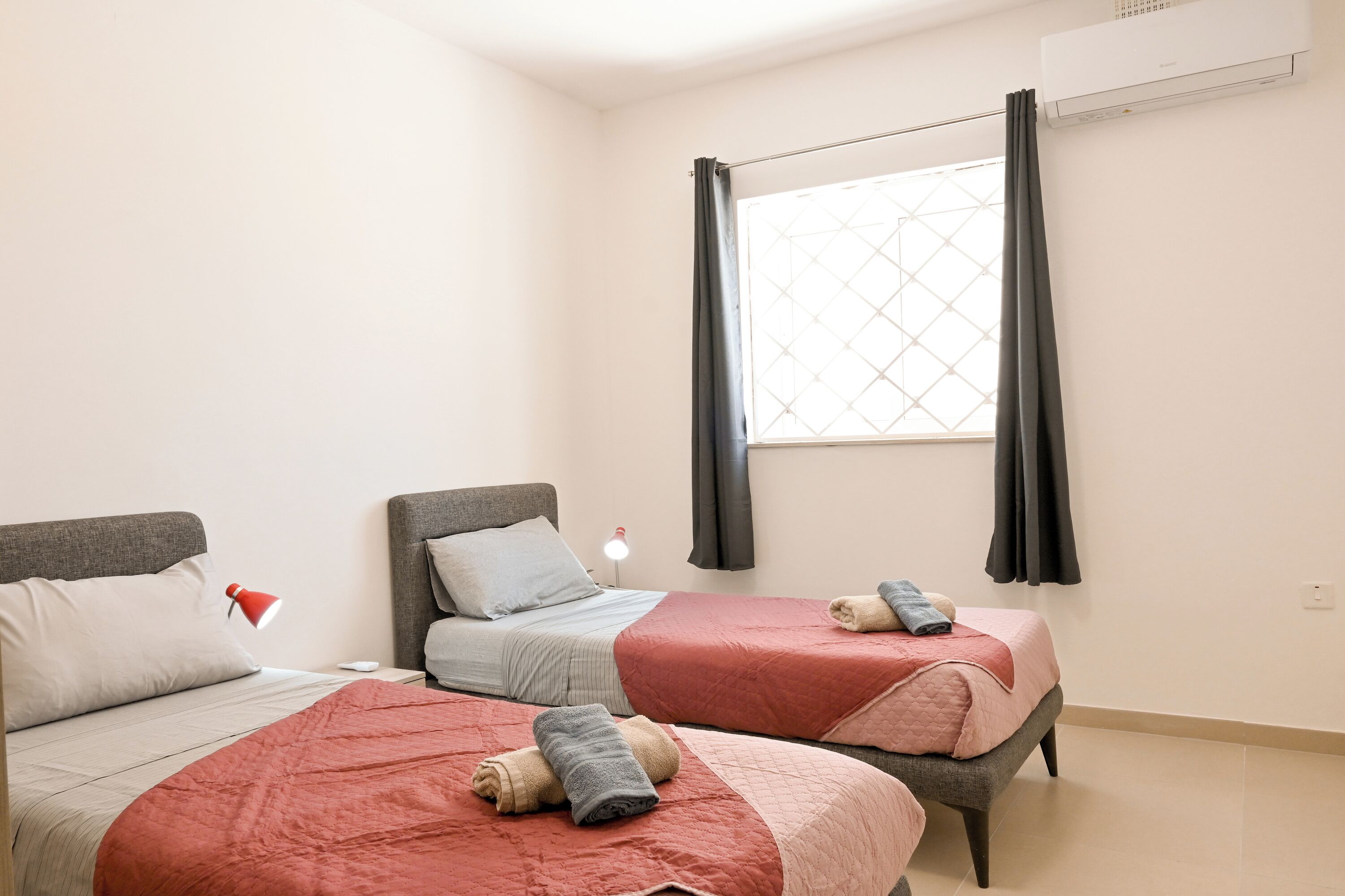 Een slaapkamer met twee bedden, een raam met gordijnen, een airconditioning en een bedlampje.