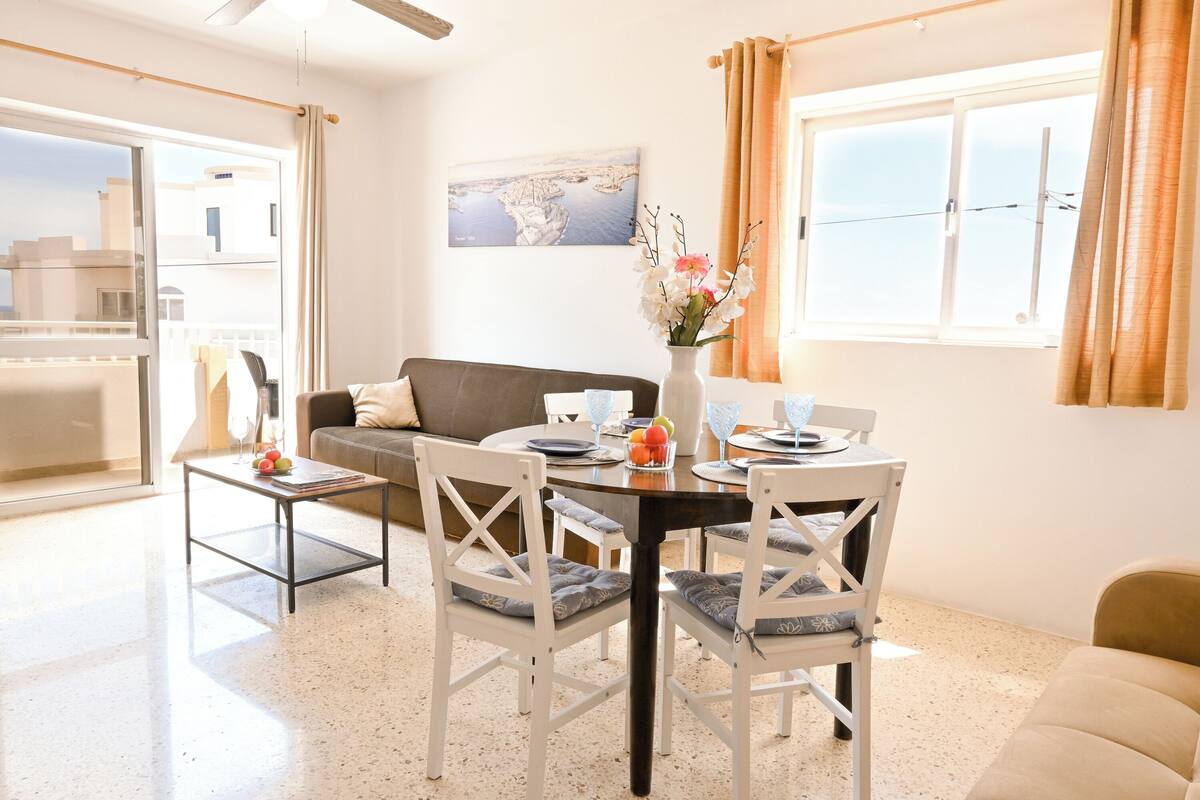 A modern living room with a dining area, white furniture, a large window with curtains, and a cityscape painting on the wall.