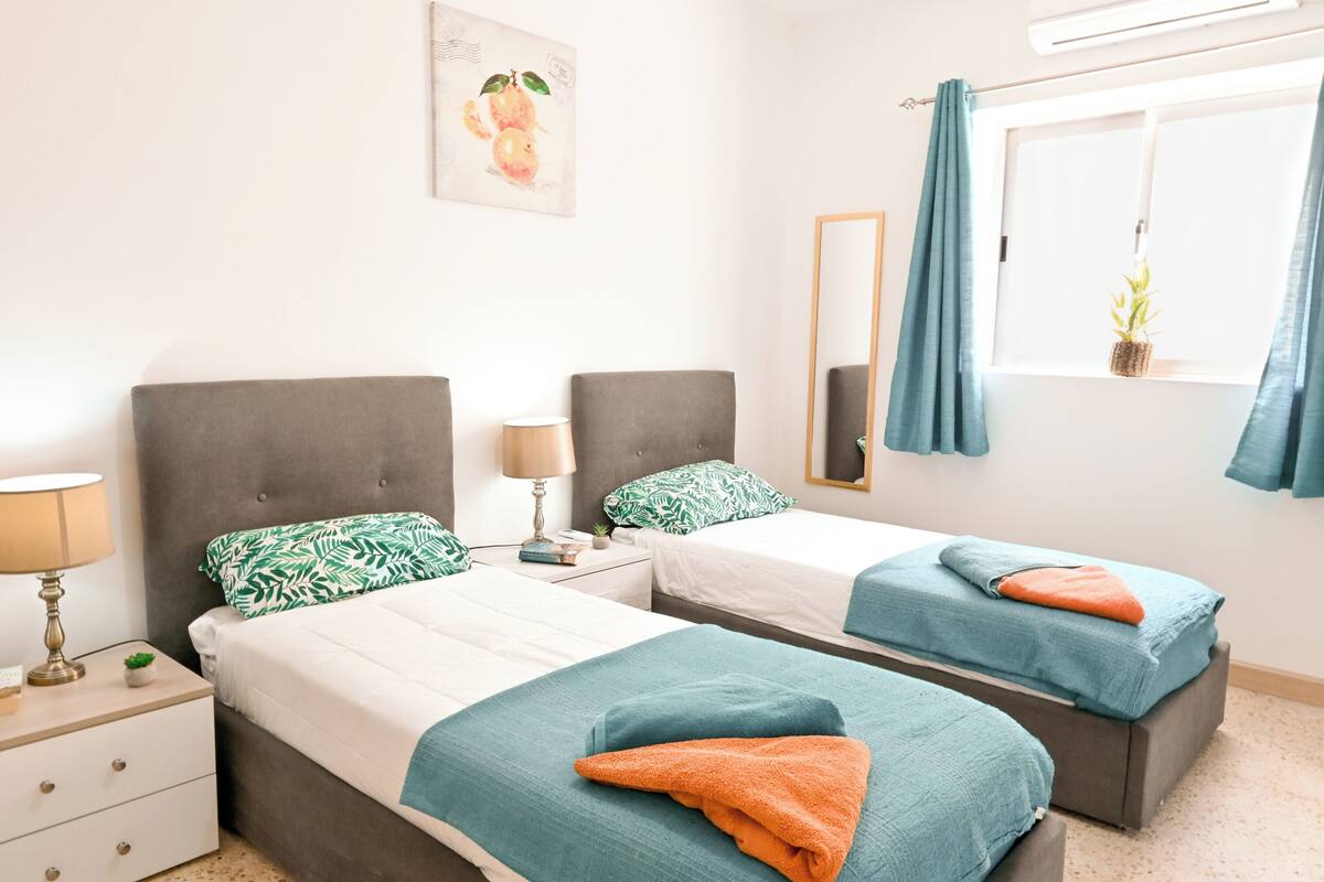 A hotel room with two beds, a nightstand, a lamp, a mirror, a window with curtains, and a wall-mounted artwork.