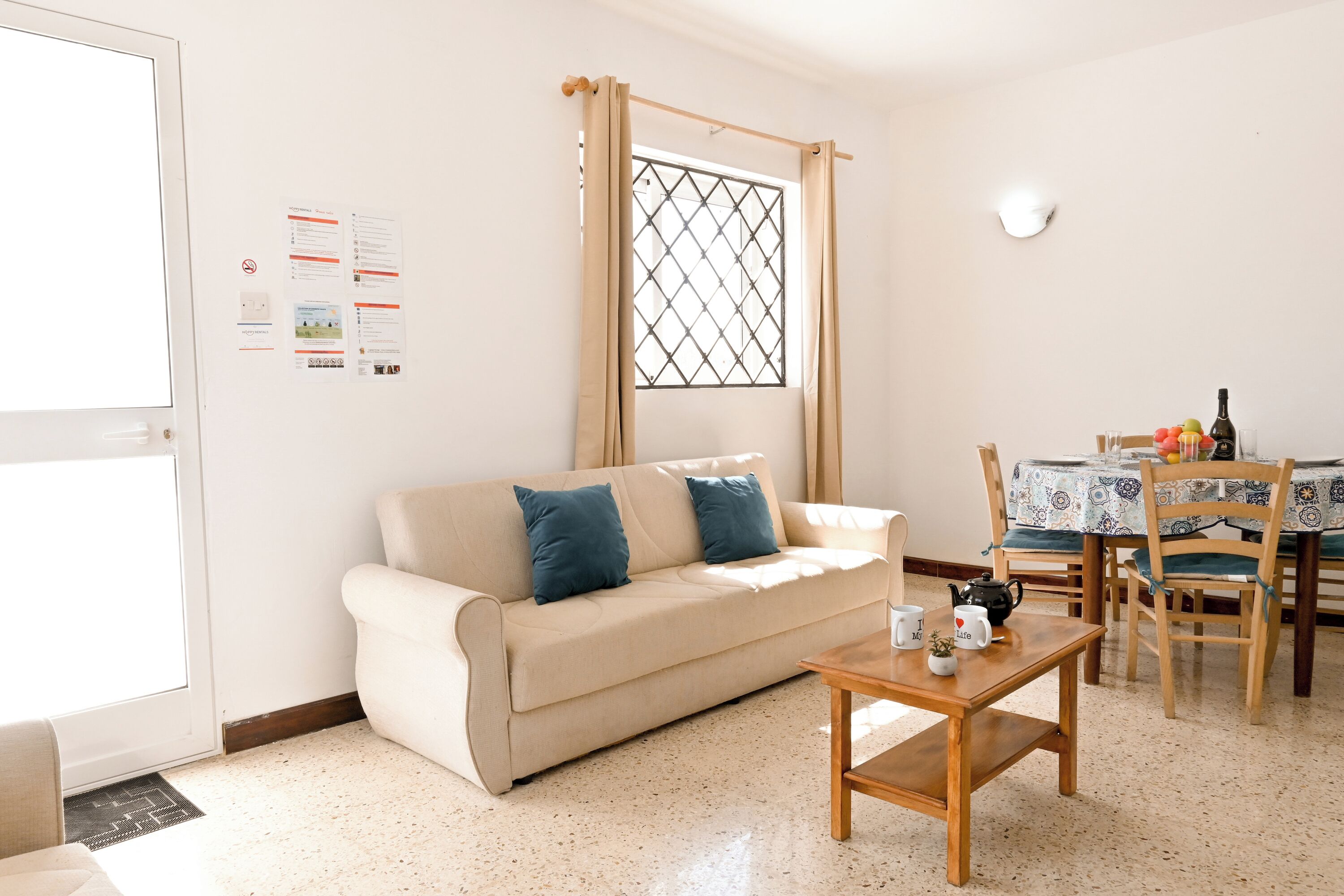A living room with a beige sofa, a wooden coffee table, a dining table with chairs, and a window with a metal grid.