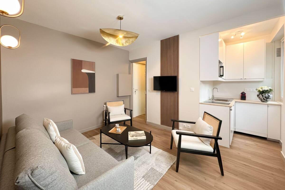A modern living room with a sofa, two chairs, a coffee table, a TV, and a kitchen area with cabinets and a sink.
