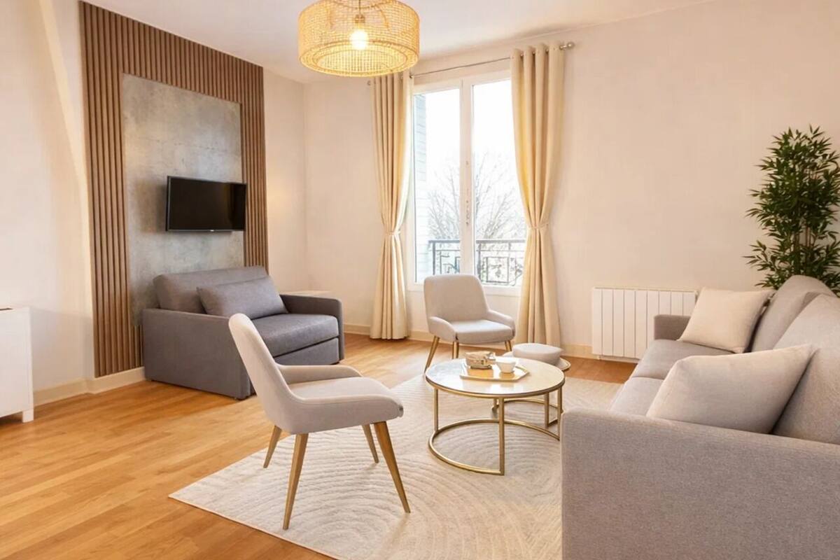 A modern living room with a sofa, armchairs, a coffee table, a TV mounted on a wall, and a large window with curtains.