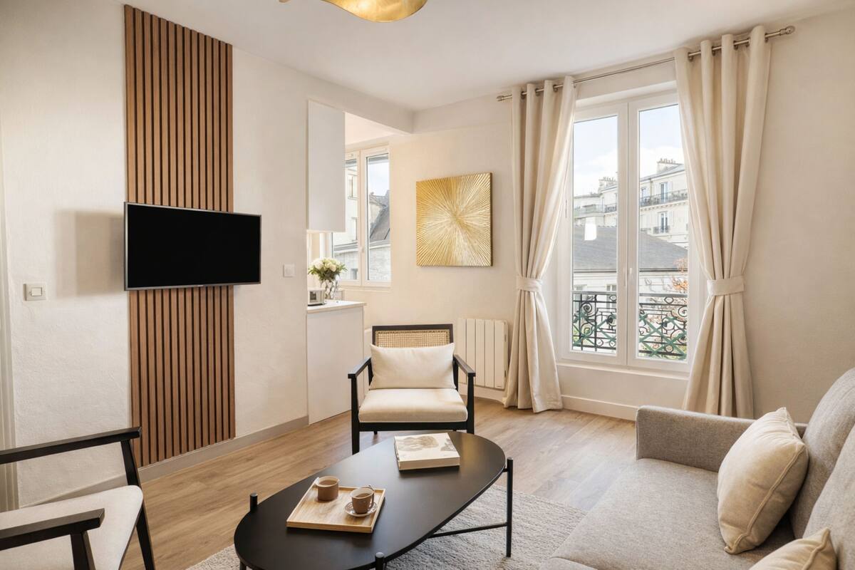 A modern living room with a flat-screen TV, a comfortable sofa, a wooden accent wall, a dining table with a tray, and a view of buildings through a window.