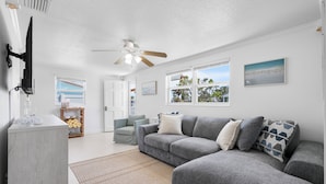 A modern living room with a grey sectional sofa, a ceiling fan, and a view of the outdoors through large windows.