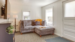A modern living room with a sectional sofa, a small side table with a lamp, and a large window providing natural light. - Swim, Hike & Unwind: Frankfort Cottage w/ Fire Pit (Frankfort)