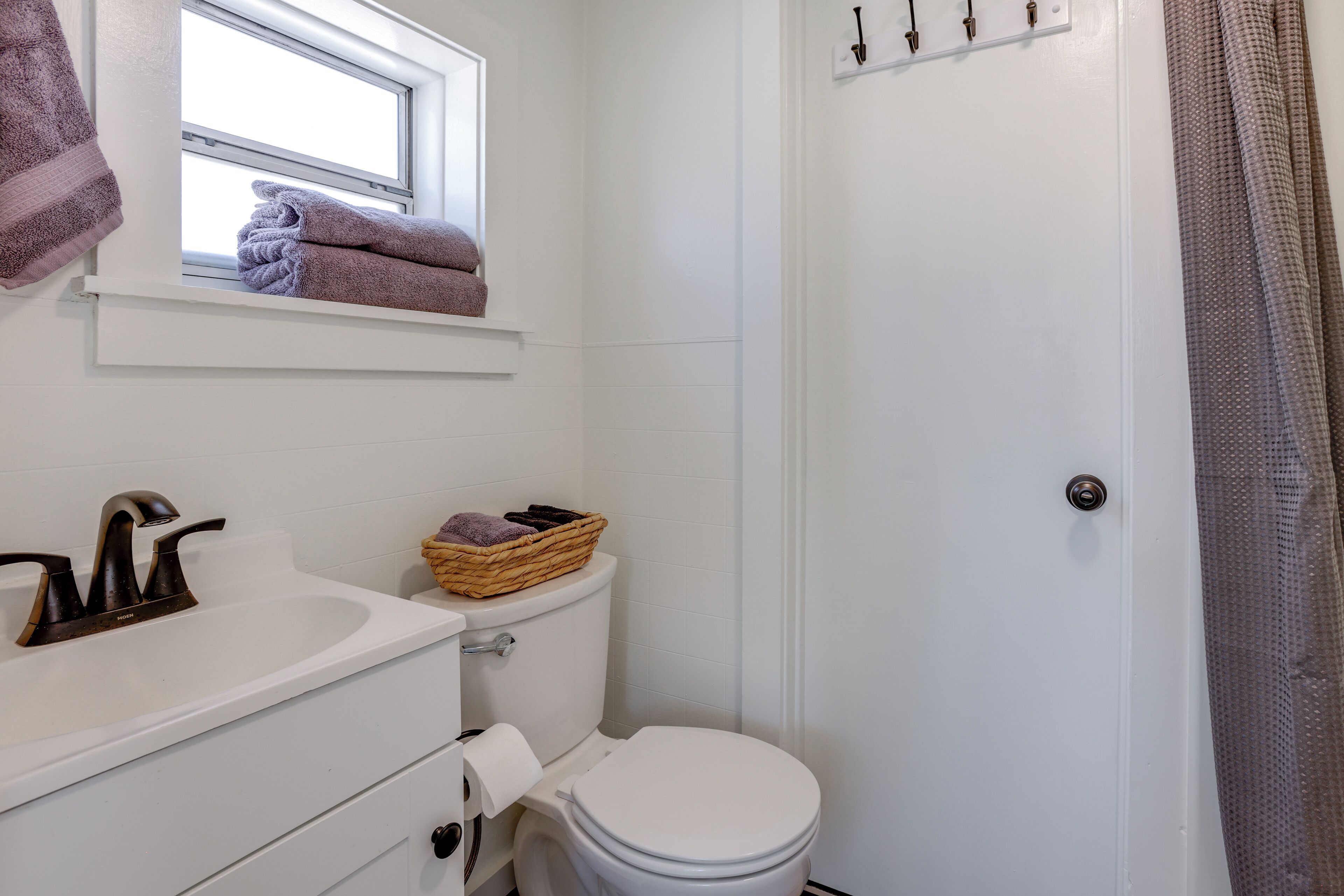 Et badeværelse med en hvid vask, toilet, et vindue med håndklæder og en bruseforhæng.