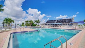A swimming pool with a clear blue water, surrounded by palm trees and a building with a solar panel roof. - Ocean Access, Mini Golf & Pools: Jensen Beach Gem (Jensen Beach)
