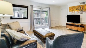 A modern living room with a leather sofa, ottoman, and armchair, a flat-screen TV on a wooden cabinet, and a large window with curtains. - Condo 9 Mi to Dtwn Dallas: BBQ By the Pool! (Dallas)