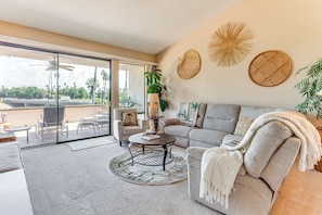 A living room with a sofa, armchair, coffee table, and a large window with a view of palm trees and a parking lot.