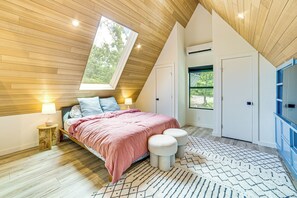 A bedroom with a sloped ceiling, a bed with a pink bedspread, two bedside tables with lamps, a patterned rug, and a view of the outdoors through a window. - A-frame Cabin w/ Deck Near Cedar Creek Lake (Malakoff)