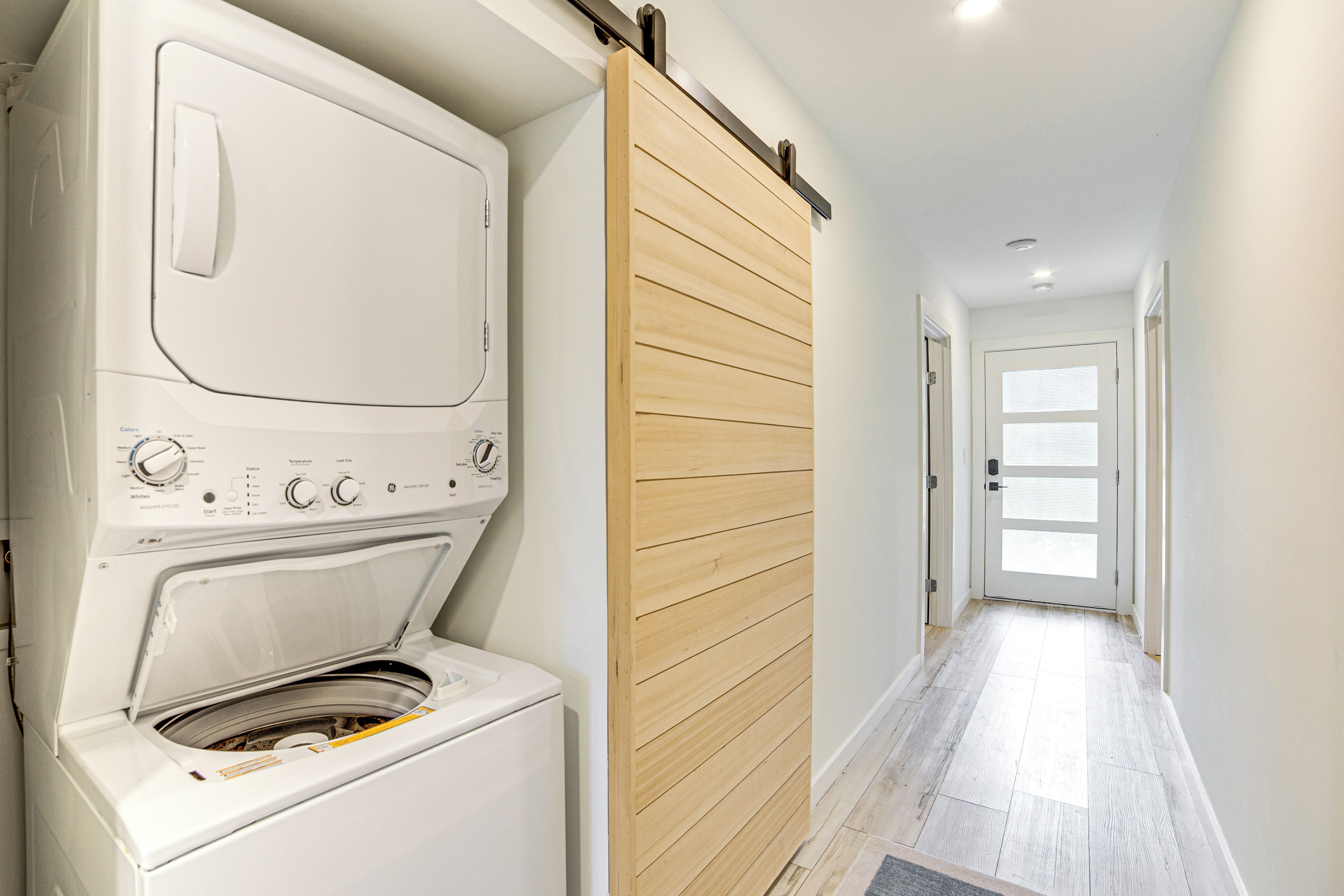A laundry room with a front-loading washer and dryer, a wooden sliding door, and a hallway leading to a door with frosted glass panels.