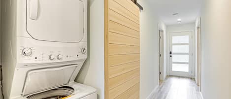 A laundry room with a front-loading washer and dryer, a wooden sliding door, and a hallway leading to a door with frosted glass panels.