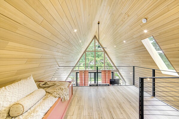 A spacious room with a sloped ceiling, a large window, a sofa, and a staircase. - A-frame Cabin w/ Deck Near Cedar Creek Lake (Malakoff)