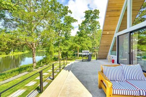 A modern outdoor patio with a wooden bench, a grill, and a glass railing overlooking a lake. - A-frame Cabin w/ Deck Near Cedar Creek Lake (Malakoff)