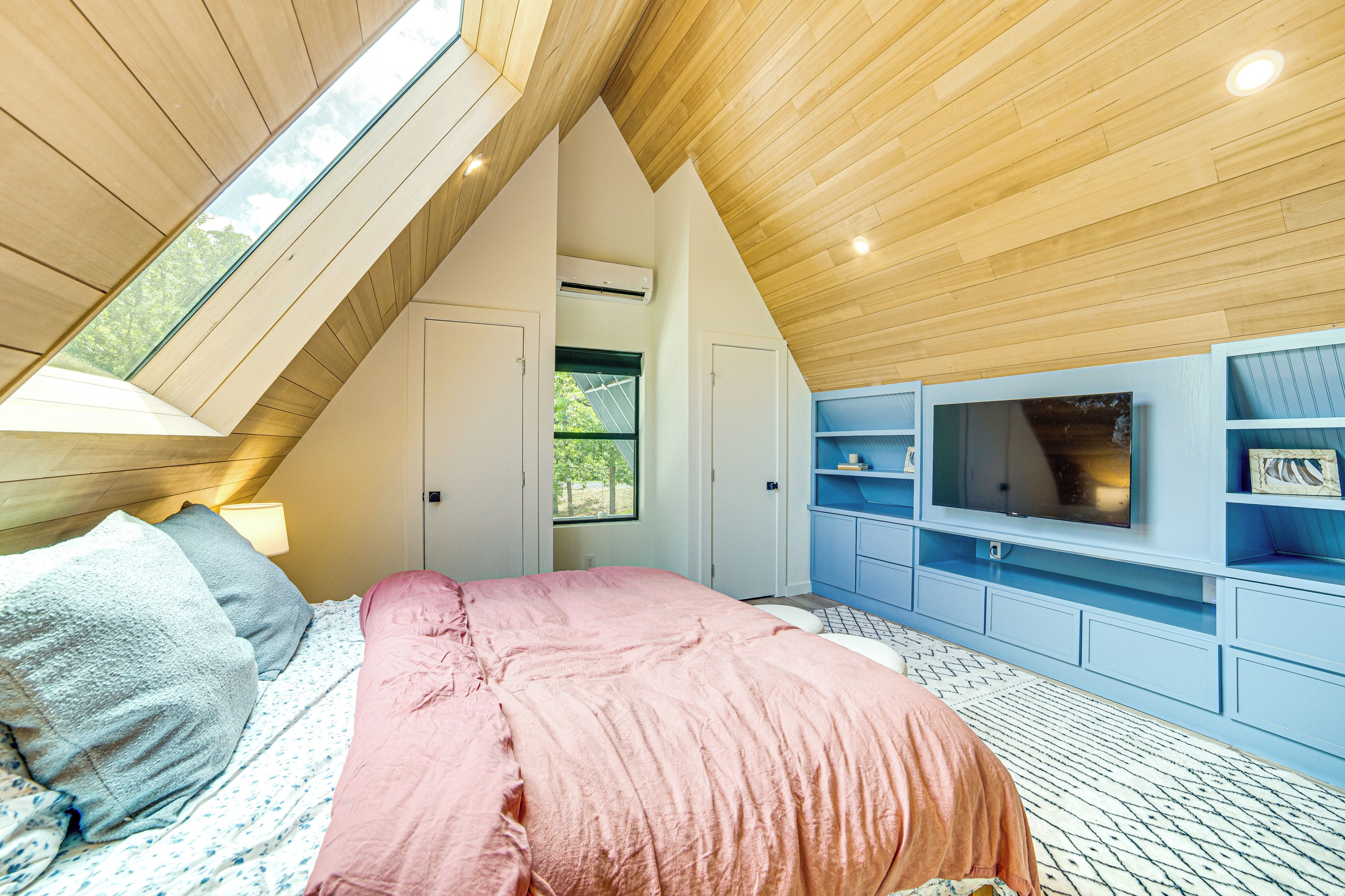 A bedroom with a sloped ceiling, a bed with pink bedding, a TV on a blue entertainment unit, and a window with a view of trees.