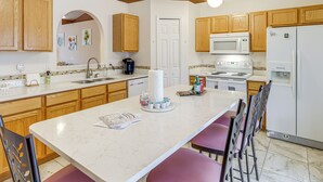 A kitchen with wooden cabinets, a white countertop, a sink, a microwave, a refrigerator, and a dining area with chairs. - Pool & Fire Pit: Family-friendly Palm Bay Home! (Palm Bay)