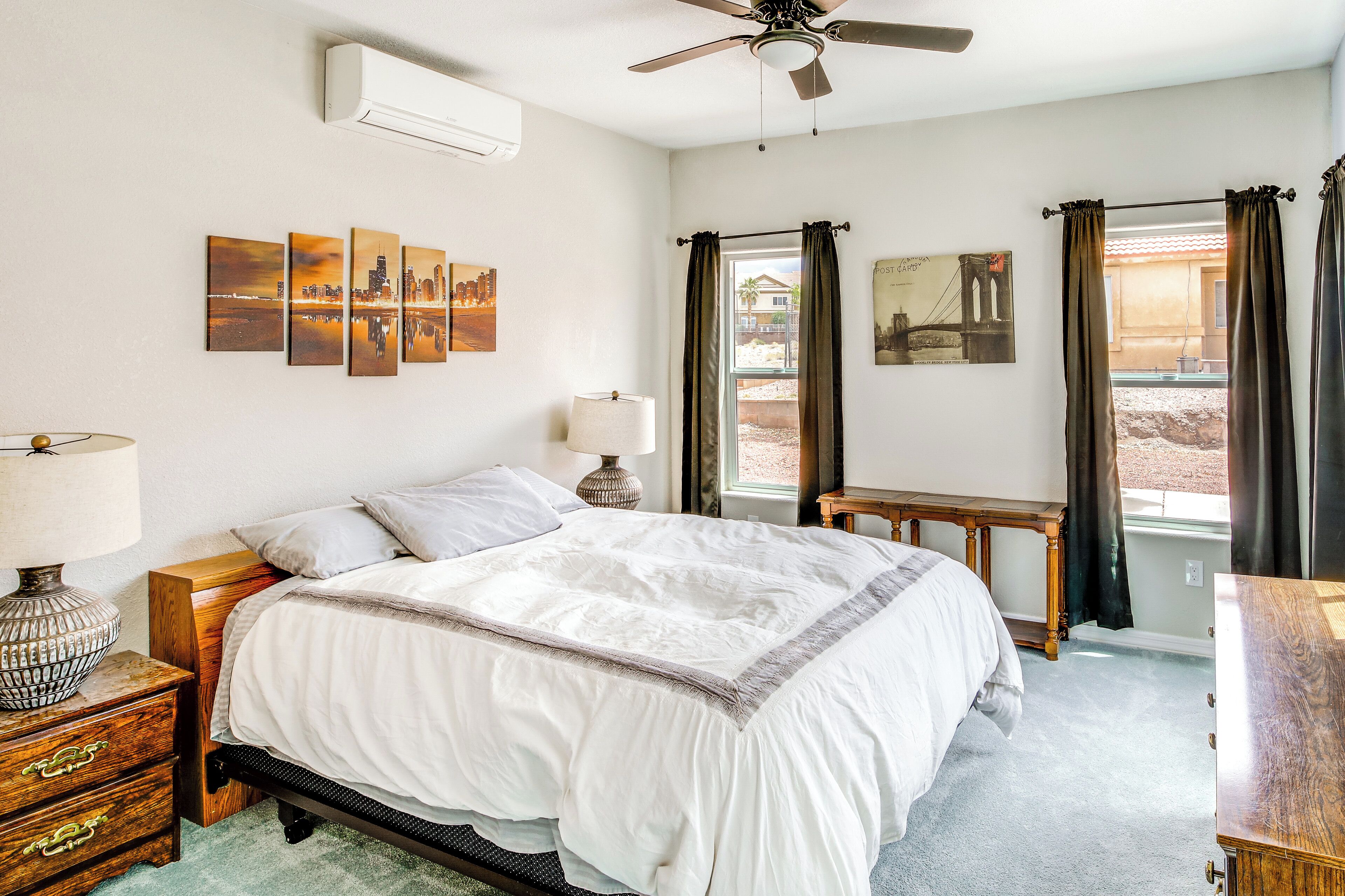Une chambre à coucher comprenant un lit, des tables de chevet, un ventilateur de plafond, des œuvres d’art accrochées au mur et une fenêtre avec des rideaux.