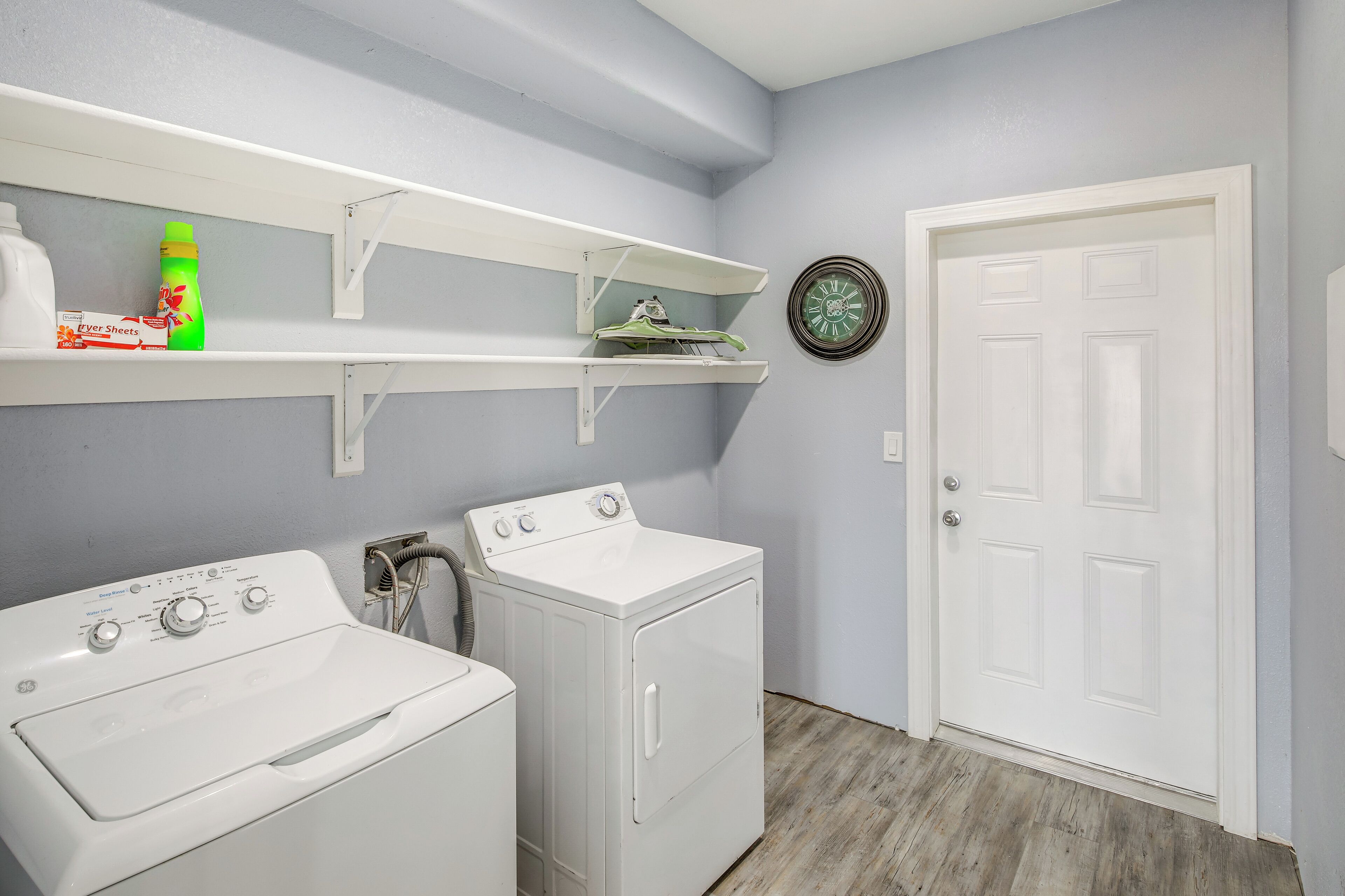 A laundry room with two white washing machines, a shelf with cleaning supplies, and a door with a clock on the wall.