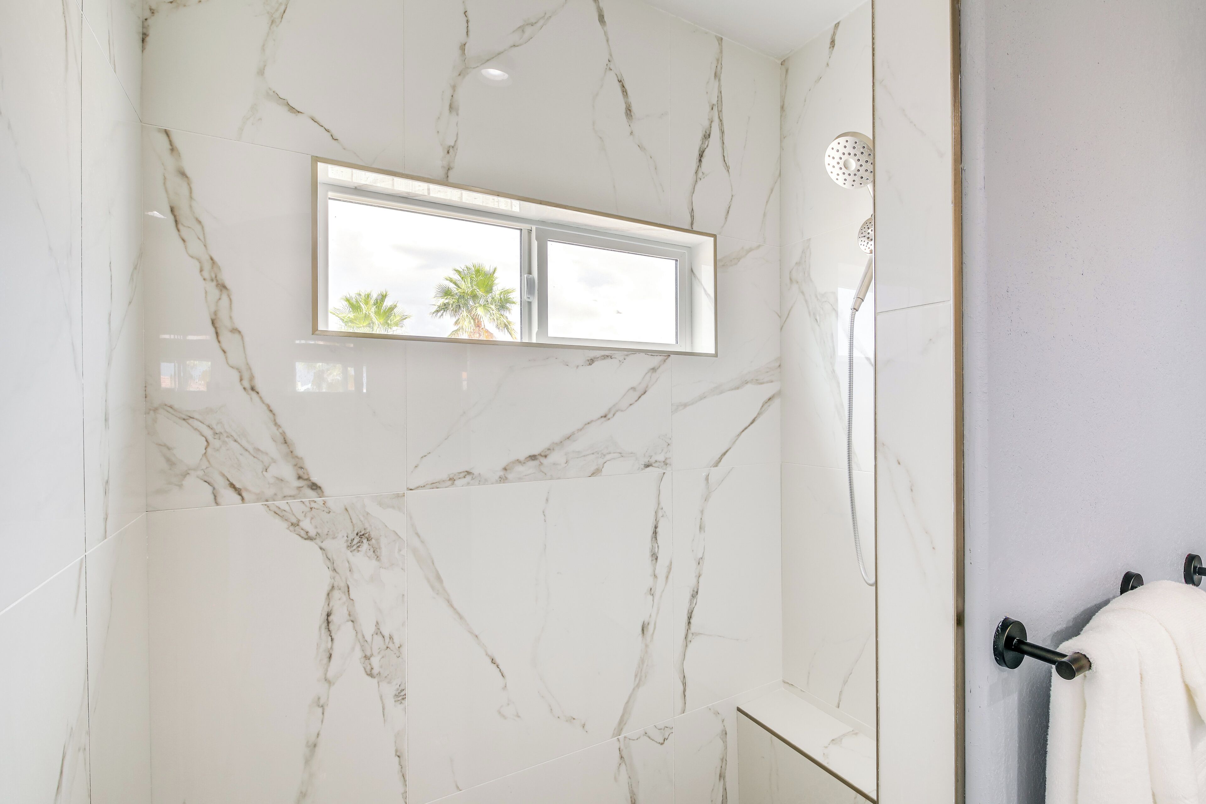 Un baño con paredes de mármol, una ventana con vistas a palmeras y un perchero.