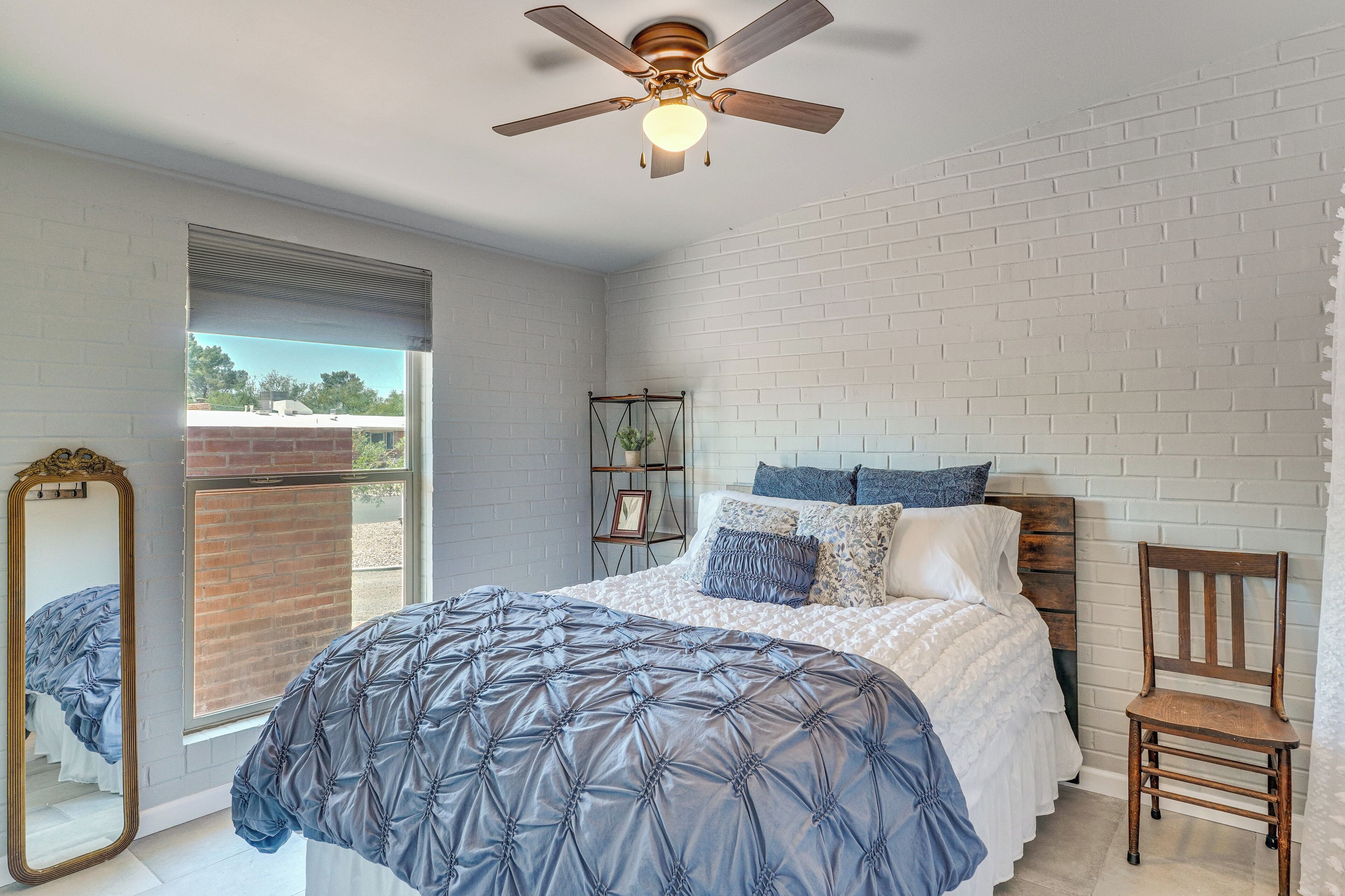 A bedroom with a bed, a wooden chair, a ceiling fan, and a window with a view of a brick wall and trees.