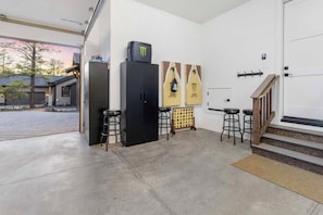 A garage with a concrete floor, a black refrigerator, a black cabinet, two bar stools, and a Monster-branded cooler on top of the cabinet.