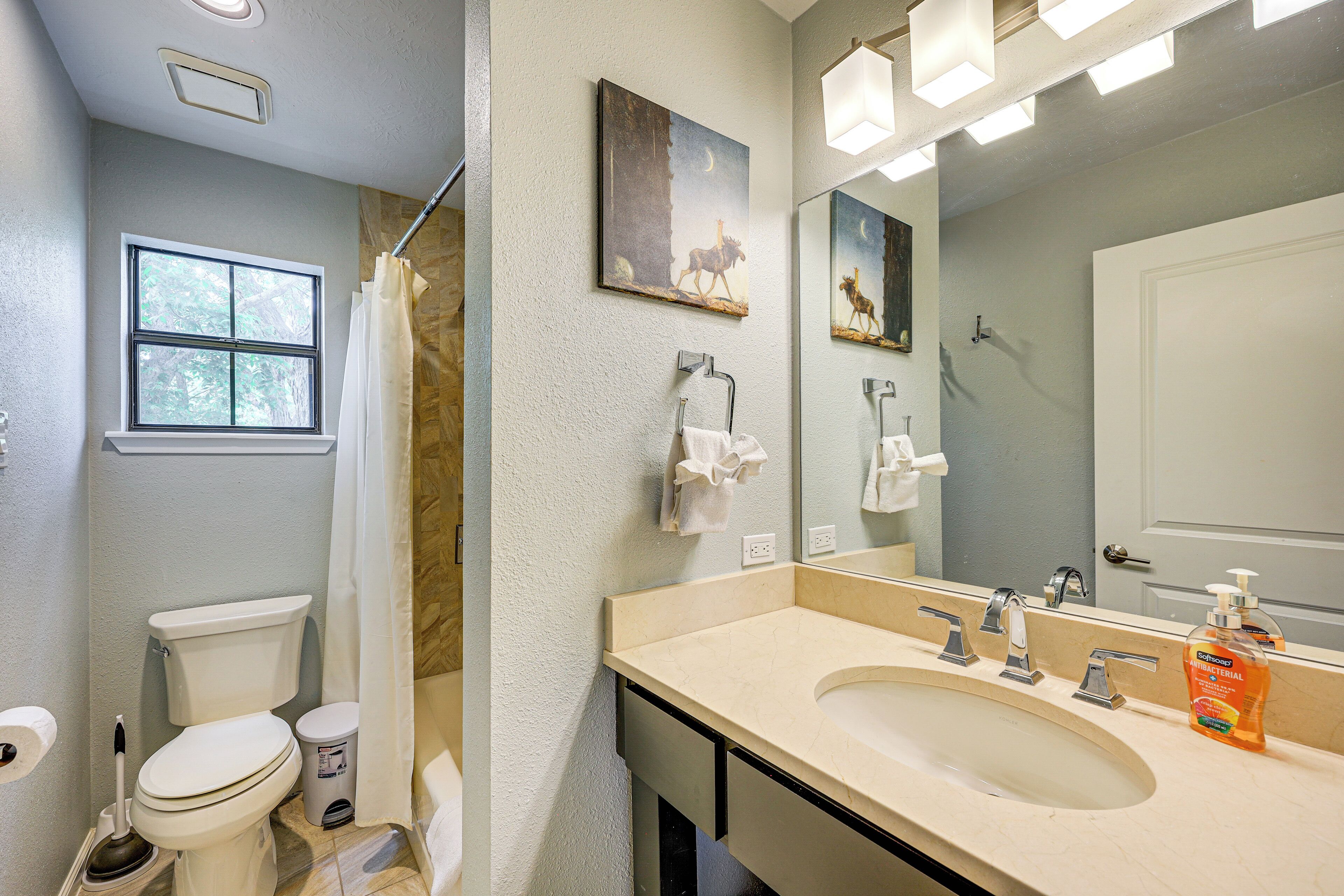A bathroom with a toilet, sink, and shower. There is a window and a painting on the wall.