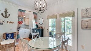 Een eetruimte met een glazen tafel en witte stoelen, een rieten stoel met een kussen met een anker, een kroonluchter, en uitzicht door de glazen deuren.