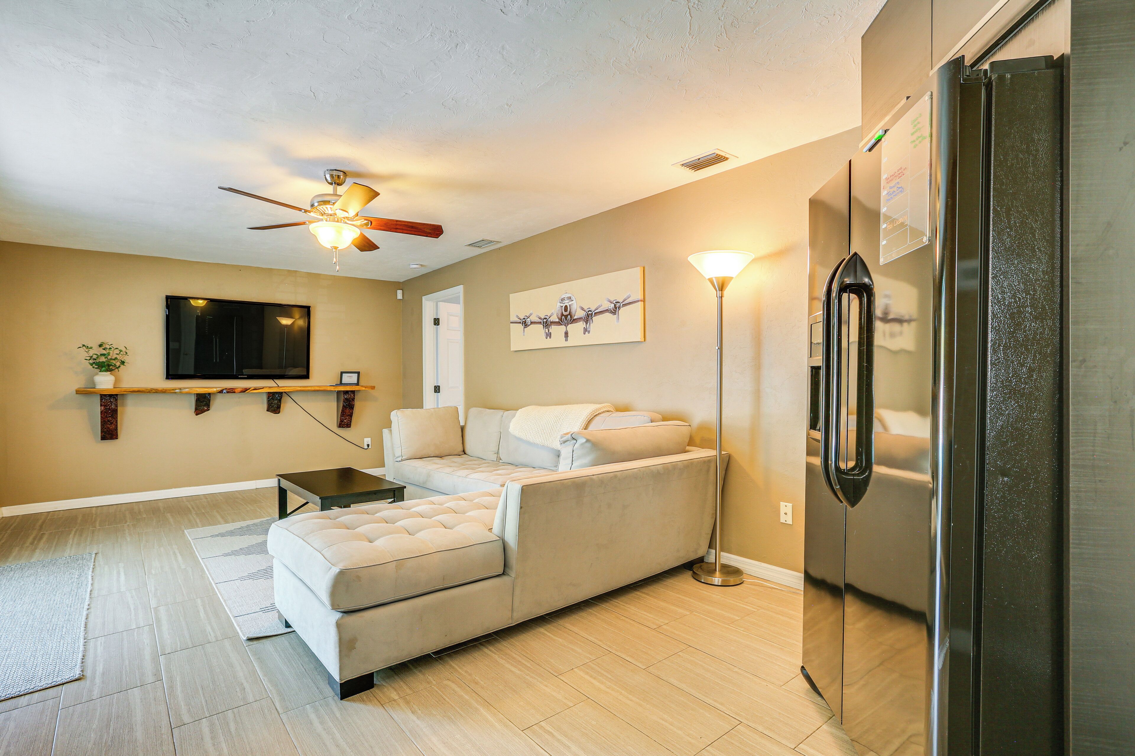 Un salon moderne avec un canapé beige, un téléviseur à écran plat, un ventilateur de plafond et un réfrigérateur en acier inoxydable.