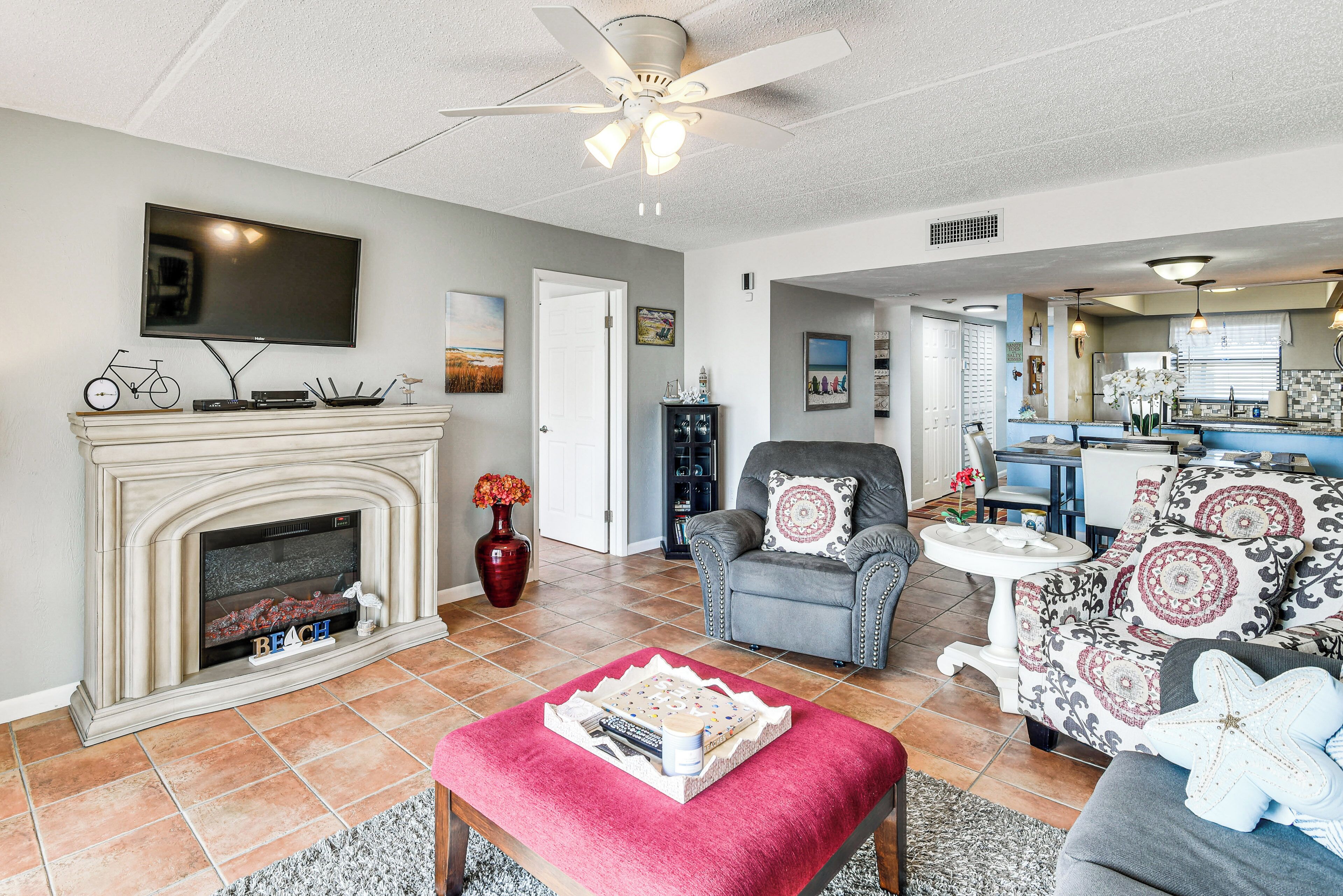 A living room with a fireplace, a television, a ceiling fan, and a dining area with a kitchen in the background.