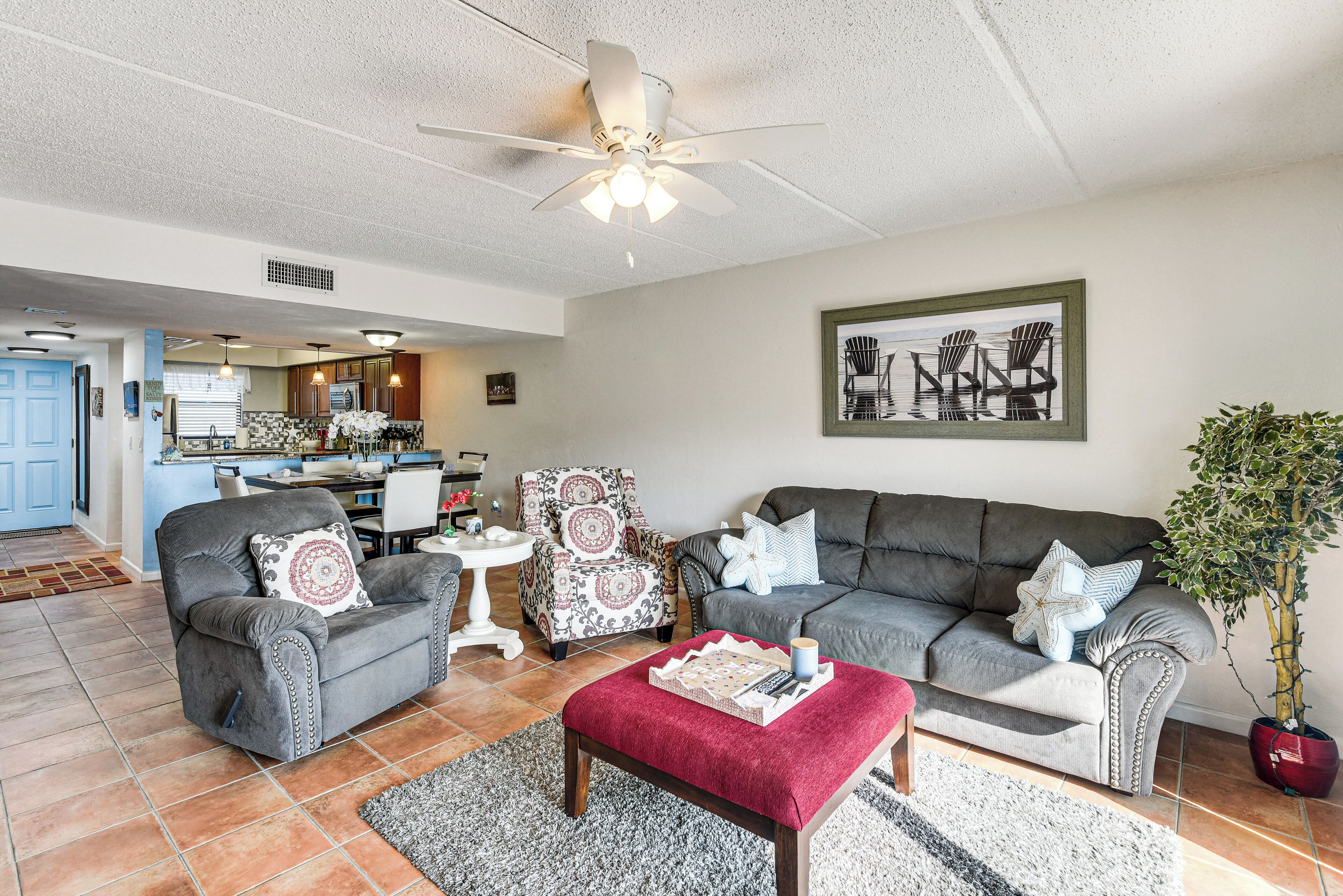 A spacious living room with a grey sofa, armchairs, and a red ottoman. There is a ceiling fan, a dining area in the background, and a potted plant.